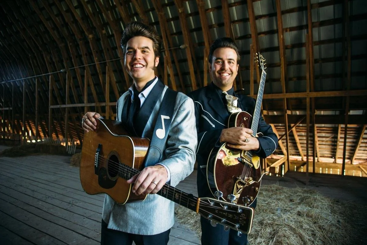 The Malpass Brothers smiling with their guitars inside a rustic barn